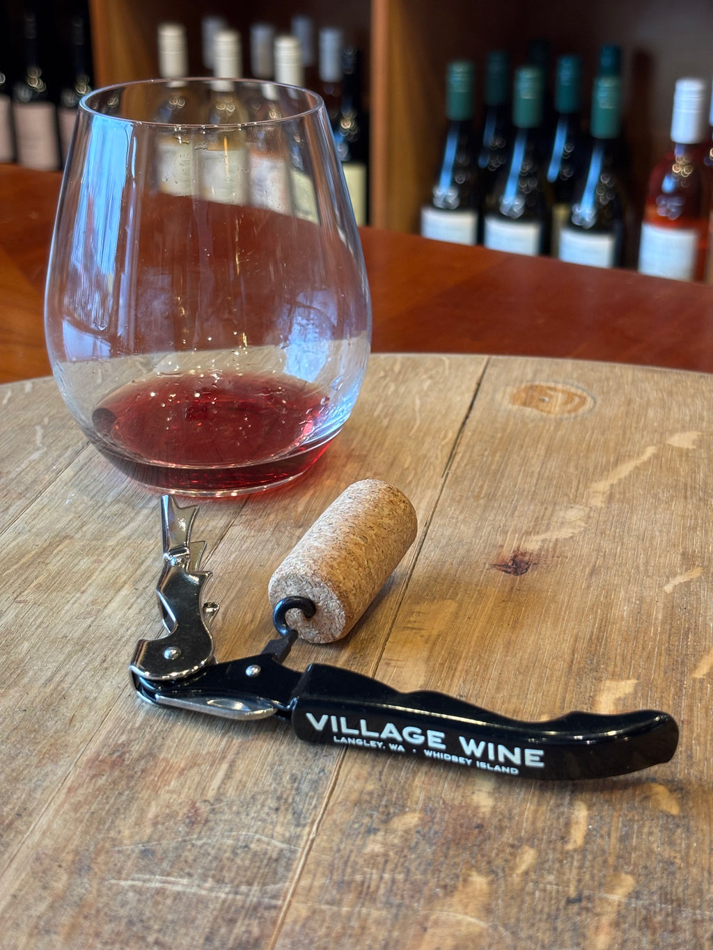 Wine glass with red wine, cork, and a corkscrew on a wooden table with wine bottles in the background.