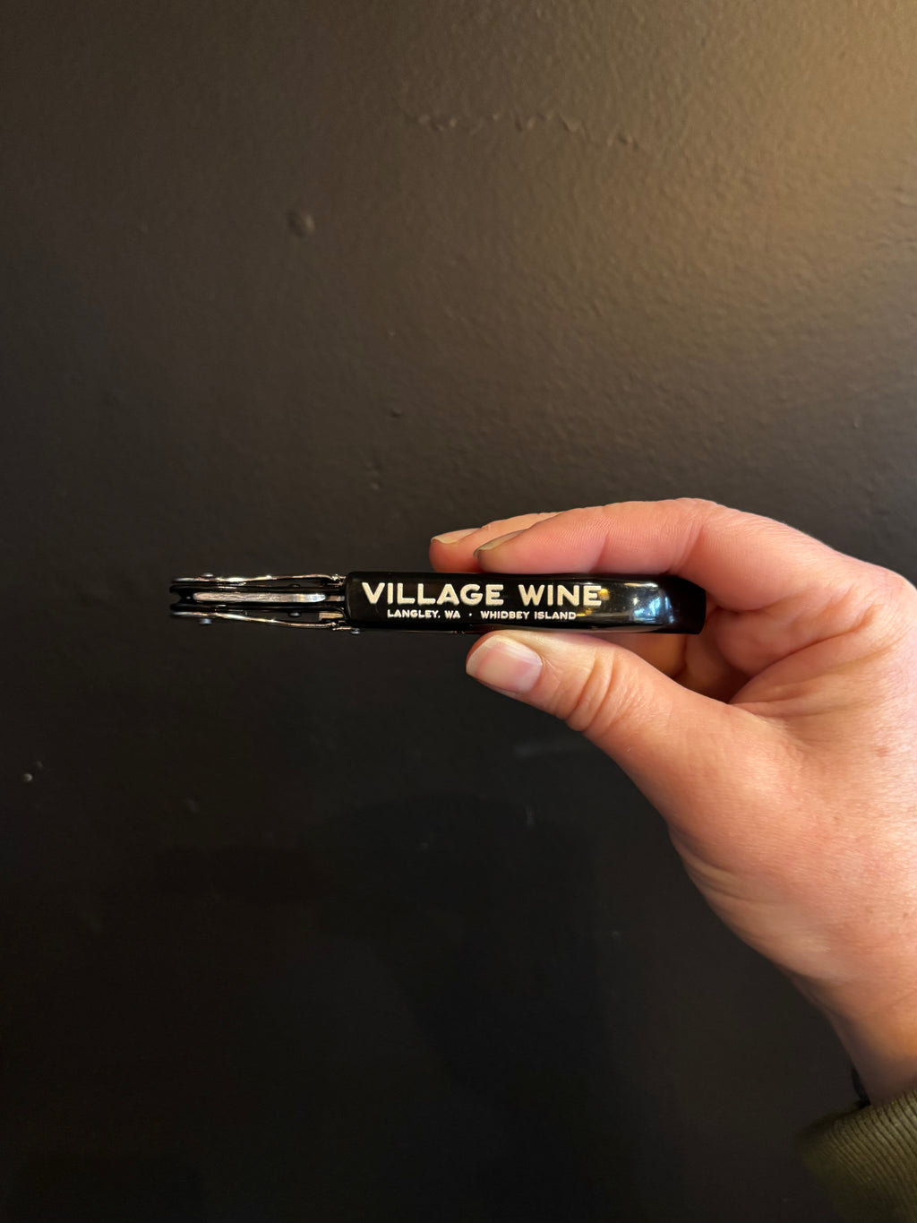 Hand holding a wine key with 'Village Wine' branding against a dark background